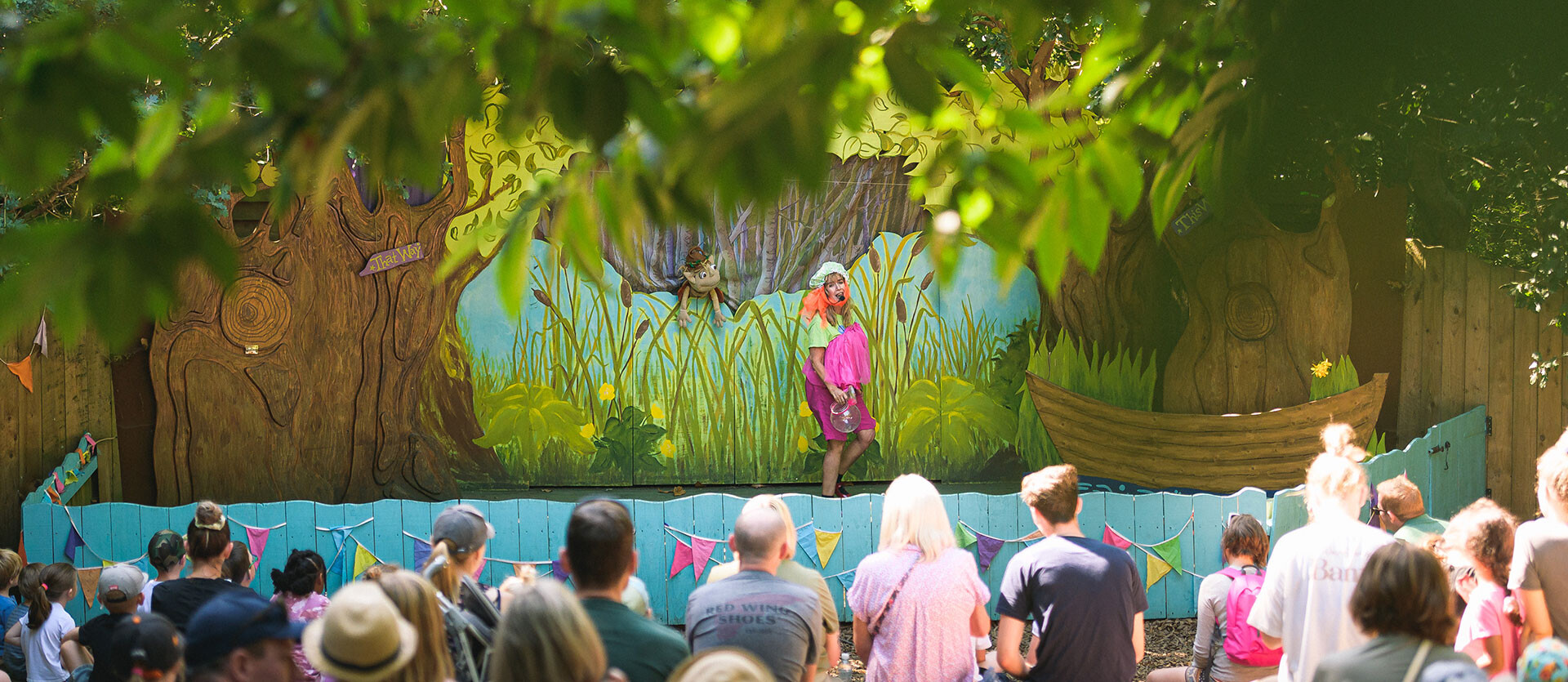 An actor and a puppet perform to a large audience on an outdoor theatre stage at BeWILDerwood Norfolk