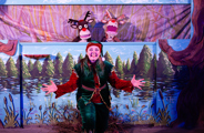 A performer dressed in an elf costume with 2 reindeer puppets behind