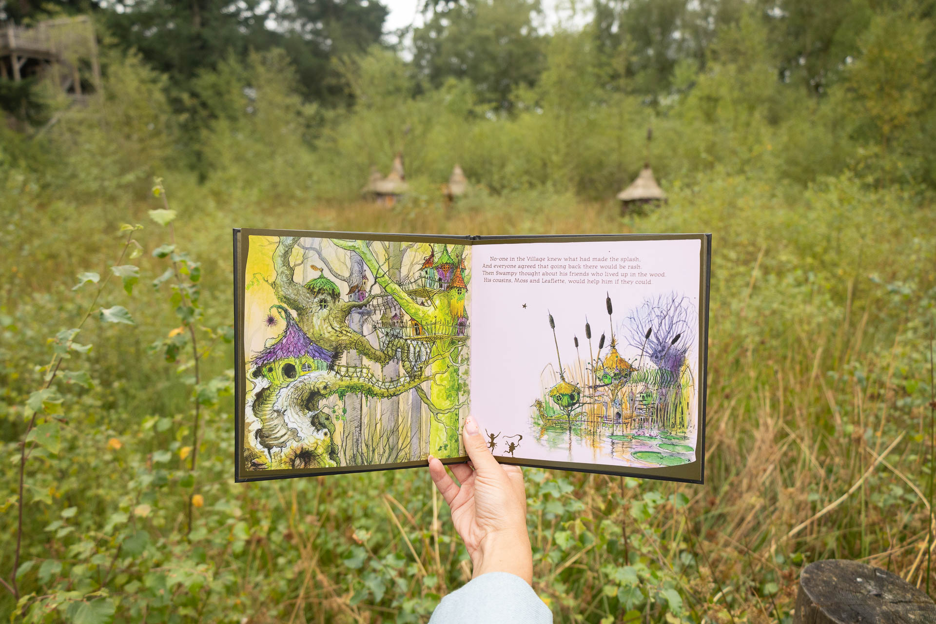 A person's hand holding a BeWILDerwood book open on a page with the Boggle Village in the background
