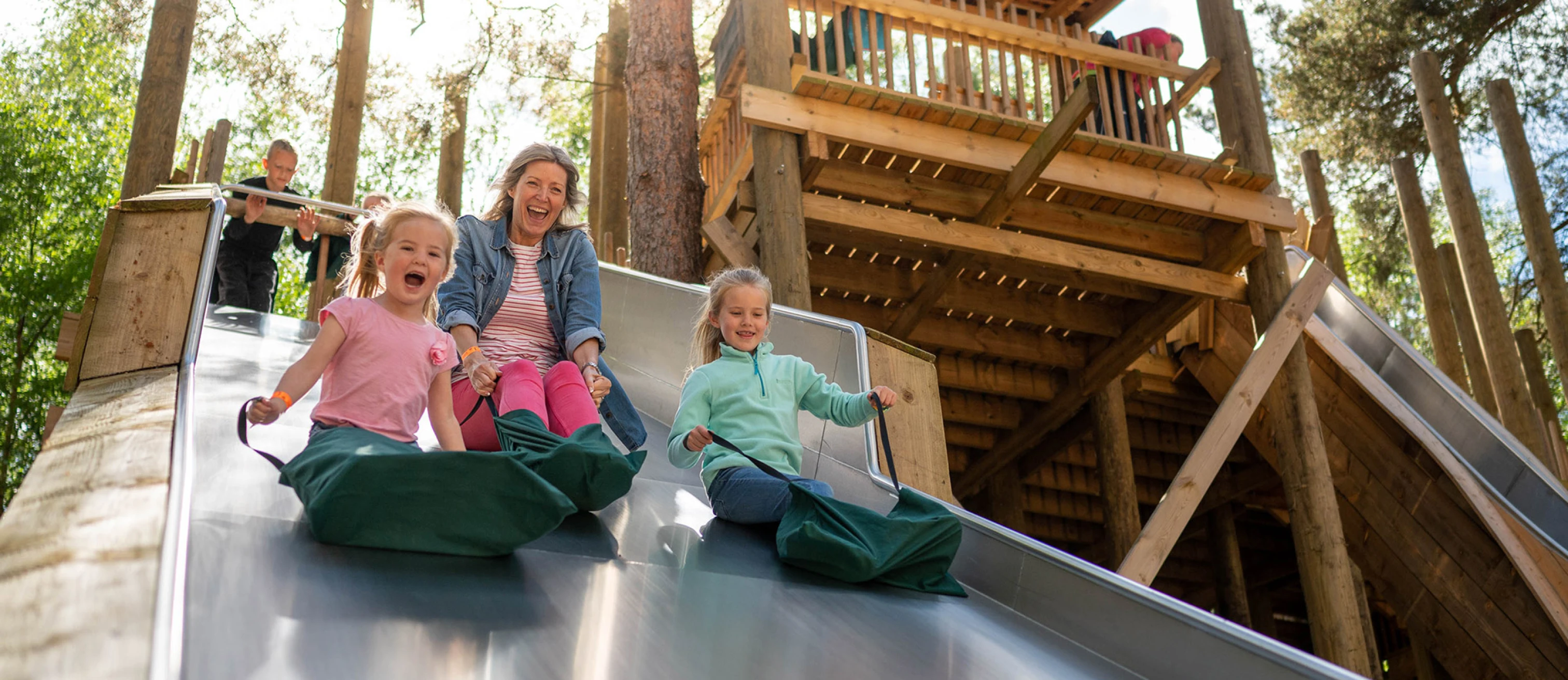 Mum and two daughters in bright colourful clothing going down a slide together at Bewilderwood