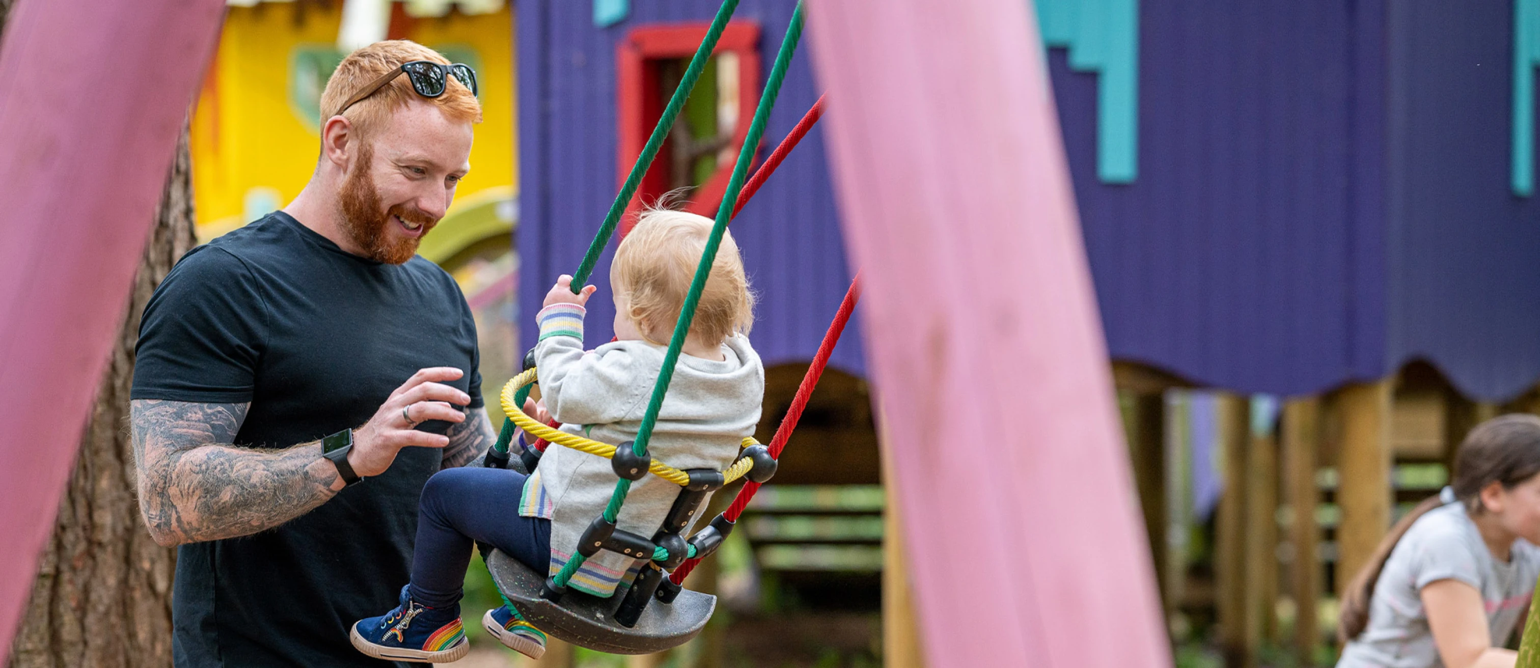 Dad pushing toddler on a baby swing with bright colourful wooden houses in the background